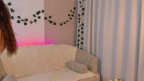 Media: A video of a cozy living room with a beige sofa adorned with a plush, white blanket and green ivy garlands. The background features a vertical striped curtain and sheer white drapes.