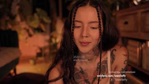 Video of a topless, tattooed, dark-haired woman with braided hair, smiling, in a cozy room with warm lighting.