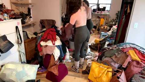 Video of a messy, cluttered living room with a woman wearing a green top and leggings, sorting through a heap of clothes and bags.