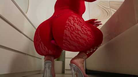 Media: Video of a curvaceous woman with large, round buttocks, wearing a tight, red, lace bodysuit, standing in a modern, minimalist room with beige walls and a large, circular mirror.