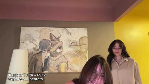 Media: Video of a woman with long dark hair and fair skin, wearing a beige shirt, standing in a room with a mustard-yellow wall, an abstract painting, and a white lamp.