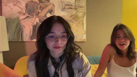 Media: Video of two young women, one with long black hair and glasses, the other with short dark hair, sitting on a couch. Background features a large, abstract painting and a yellow wall.