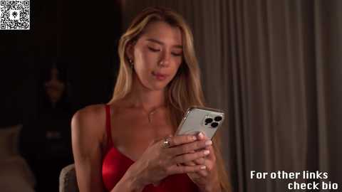 Video of a blonde woman with long hair, wearing a red dress, looking at a smartphone, in a dimly lit room with a QR code in the corner.