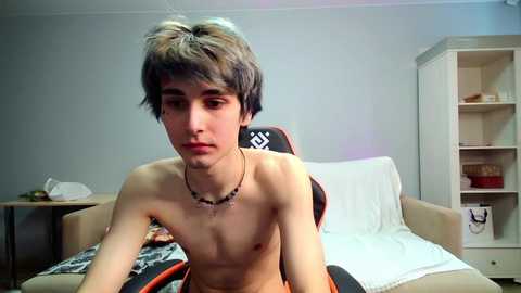 Video of a young, thin, shirtless Caucasian male with short, dark hair, sitting on a gaming chair in a sparsely furnished bedroom. Background includes a bed with white sheets, beige walls, and a white bookshelf.