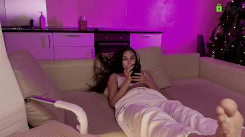 A video of a young woman with long black hair, wearing a white tank top and pajama pants, lying on a beige sofa, holding a phone to her mouth, in a dimly lit room with purple lighting, kitchen cabinets in the background.