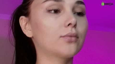 Video of a young woman with fair skin and long dark hair, wearing makeup, against a purple background. She has a neutral expression and visible ear piercing.