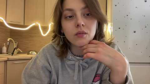 Video of a young girl with light skin, shoulder-length brown hair, and wearing a gray hoodie, leaning against a kitchen counter with wooden cabinets and a glowing neon light strip.