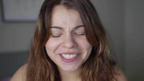 Media: Video of a smiling young woman with wavy brown hair, fair skin, and closed eyes, set against a blurred, neutral background.