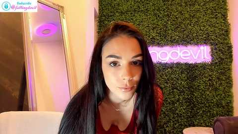 Media: Video of a young woman with long black hair, fair skin, and a red top, standing in front of a green wall with a neon \"modevill\" sign.