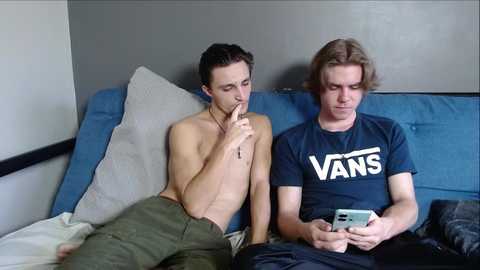 Video of two shirtless men on a blue couch, one holding a finger to his lips, the other reading a phone.