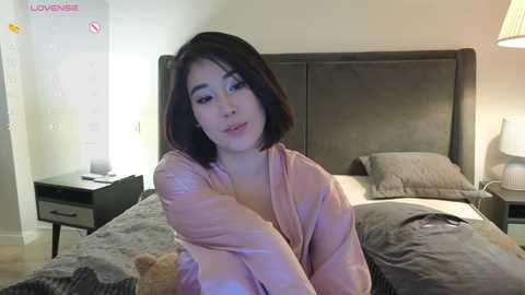 A video of a young Asian woman with shoulder-length dark hair, wearing a light pink robe, sitting on a bed with gray bedding in a modern bedroom.