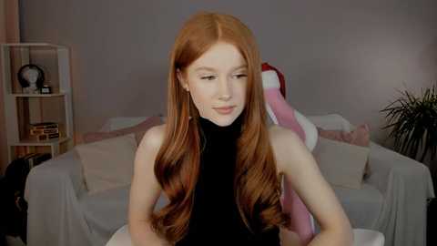 Media: Video of a young, fair-skinned, red-haired woman with long, wavy hair, wearing a black turtleneck, sitting in a cozy, softly lit room with white furniture, a bookshelf, and a potted plant.