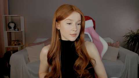 Video of a fair-skinned, long-haired redhead woman in a black turtleneck, seated in a living room with beige furniture and a pink plush toy, against a muted grey wall.