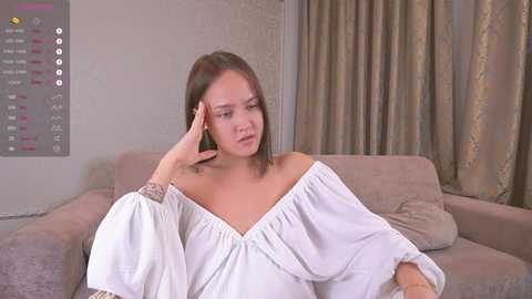 A video shows a young Asian woman with straight brown hair, wearing a white off-shoulder blouse, sitting on a beige couch, with a distressed expression, touching her head, in a modern living room with beige walls and curtains.