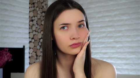 Video of a young, fair-skinned woman with straight, dark brown hair and blue eyes, resting her chin on her hand, wearing a strapless top, in a modern room with wooden walls and a pink floral decoration.