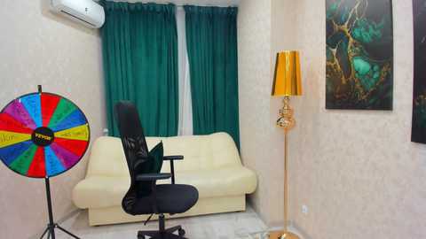 Video of a minimalist room with a cream sofa, black office chair, teal curtains, a colorful game spinner, a tall gold lamp, and an abstract painting on beige walls.