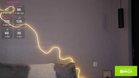 Video of a dimly-lit room with a gray wall, a bed with white and gray pillows, a glowing yellow wire looped around the room, and a black pendant light on the right.