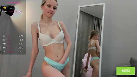 Video of a slim, blonde Caucasian woman in white lace bra and light blue panties, standing in front of a mirror, in a modern bedroom with gray walls and a white door.