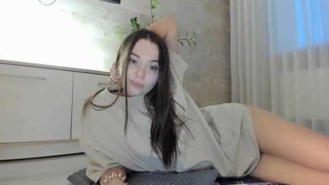 Media: A video of a young woman with long brown hair, fair skin, and a slender physique, lying on her side in a light gray t-shirt, in a modern, minimally decorated room with white walls and a wooden floor.