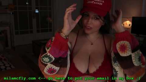 Media: Video of a curvy woman with large breasts in a red knit sweater, red hat, and red bra, adjusting her hat in a cozy, dimly-lit living room.