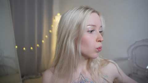 Media: Video of a fair-skinned, blonde woman with long hair, wearing a sheer, floral-patterned top, blowing a kiss in a softly lit, minimalistic room with beige walls and fairy lights.