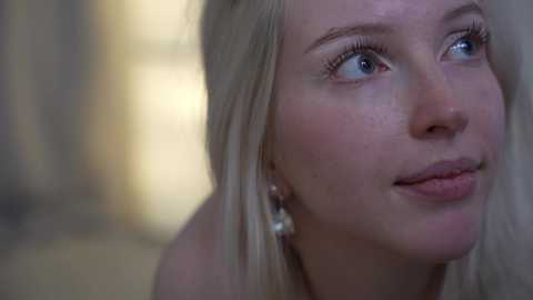 A close-up video of a fair-skinned woman with long, straight blonde hair, wearing a pink lip gloss, and light makeup. She has blue eyes and is looking upwards, slightly smiling. The background is blurred, featuring neutral tones.