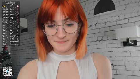 Video of a fair-skinned woman with shoulder-length, vibrant orange hair, wearing glasses and a sleeveless white top, standing in a modern, white-brick-walled room.