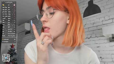 Media: Video of a pale-skinned woman with orange hair, wearing round glasses, a white t-shirt, and a finger to her mouth, in a modern, white-brick-walled room.