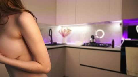 Video of a nude woman with light skin and small breasts, standing in a modern kitchen with white cabinets, purple LED lights, and a round ring light above the stove.