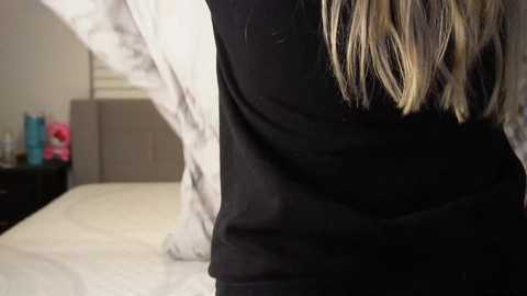 Video of a person with long blonde hair, wearing a black shirt, standing on a bed in a dimly lit bedroom.