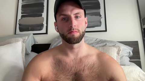 Video of a shirtless, bearded, light-skinned man with a trimmed beard and a black baseball cap, lying on a bed with white pillows and gray bedding.