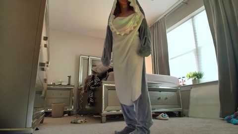 Media: Video of a woman in a light blue, hooded onesie standing in a cluttered bedroom with a mirror, dresser, and bed.