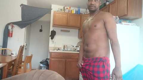 Video of a shirtless Black man with a small build, wearing red and black plaid shorts, standing in a cluttered, modest kitchen with wooden cabinets, a fridge, and a stove.