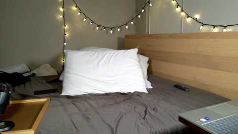 Video of a cozy, minimalistic bedroom with a light wooden headboard, white pillows, and a gray duvet. Warm, string lights hang above the bed, creating a soft ambiance. A laptop and remote control are placed on the bedside table.