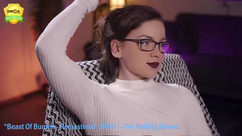 Media: Video of a young woman with light skin, brown hair, and glasses, wearing a white ribbed sweater, reclining in a black and white patterned chair. She has a confident expression.