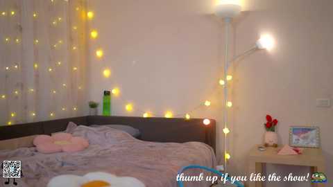 Video of a cozy bedroom with a gray bed, white walls, fairy lights, a standing lamp, and a QR code in the corner.