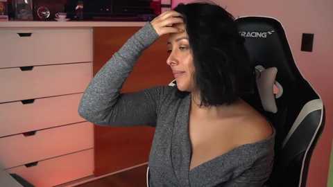 Video of a light-skinned woman with shoulder-length black hair, wearing a gray off-shoulder top, sitting in a black gaming chair, scratching her head. Background features a white dresser and pink walls.