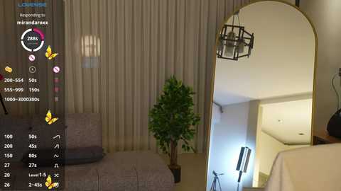 Video of a modern living room with beige curtains, a large mirror, a brown sofa, a small potted plant, and a sleek black chandelier. The room is warmly lit.