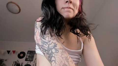 Video of a young woman with long, wavy black hair and pale skin, wearing a white tank top and a colorful, floral sleeve tattoo, standing in a room with posters and a record player on the wall.