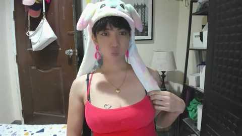 Media: Video of a woman with fair skin, short dark hair, wearing a pink tank top, pink earrings, and a bunny hat, in a cozy, cluttered room with a white purse hanging, a lamp, and a bookshelf.