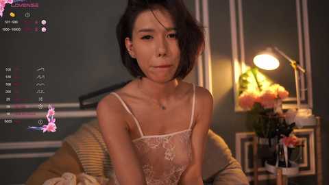 Media: Video of a young Asian woman with short black hair, wearing a sheer lace bra, sitting on a bed with a warm yellow lamp and pink flowers in the background.