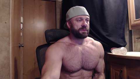 Video of a muscular, shirtless man with a beard, wearing a backwards baseball cap, sitting in a chair in a dimly lit room with wooden walls and a black curtain.