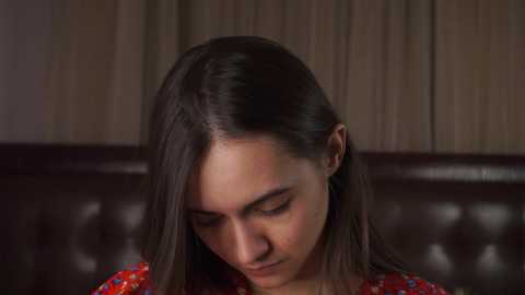 A close-up video of a young woman with straight, dark brown hair, wearing a red-patterned top, sitting on a dark leather couch. The background features beige curtains.