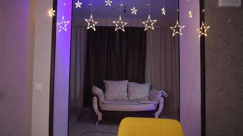 Video of a cozy living room with a white couch, yellow chair, star-shaped string lights, and a black curtain backdrop.