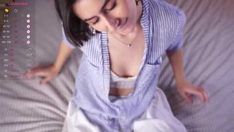 Video of a young woman with short dark hair, wearing a white bra and a blue and white striped cardigan, lying on a bed with a gray blanket.