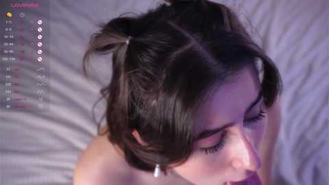 Video of a young woman with brown hair in a messy bun, lying on a white bed, eyes closed, possibly asleep, with a digital clock displaying 3:14 AM in the top left corner.