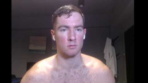 Media: A video of a shirtless, fair-skinned man with short, dark hair, wearing white earbuds, in a dimly lit, sparsely decorated room with a TV and a white robe hanging on a hook.