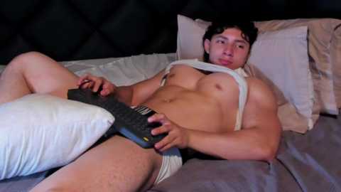 Media: Video of a muscular, light-skinned man with dark hair, lying on a bed in underwear, holding a remote, shirt partially off, against a dark, tufted headboard.