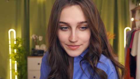 Media: Video of a young woman with fair skin and long brown hair, wearing a blue shirt, smiling gently. Background features green curtains, a plant, and glowing neon lights, creating a cozy, modern indoor setting.