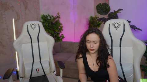 Video of a woman with curly dark hair in a black top, seated in a white gaming chair with tribal patterns, surrounded by green plants and purple lighting in a modern room.